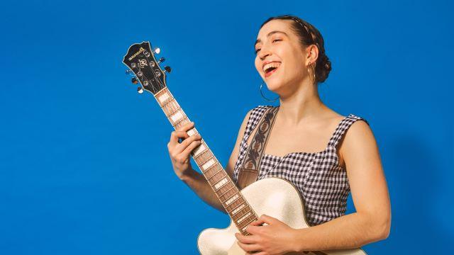 Alisa Amador smiles while singing and playing guitar. 她穿着格子布连衣裙，站在明亮的蓝色背景前.