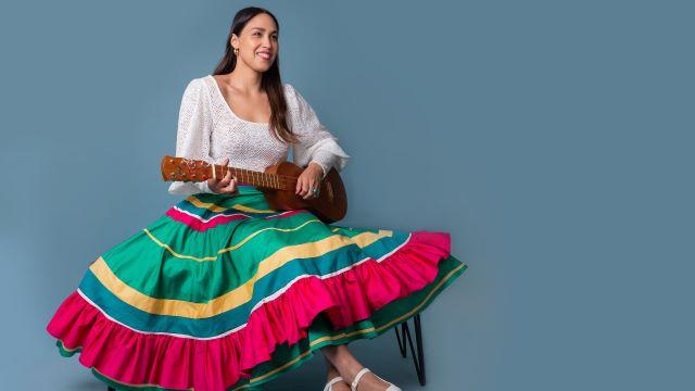 Sonia De Los Santos sits on a chair in 一个大的, striped, colorful skirt. She is holding a guitar-like instrument and smiling.